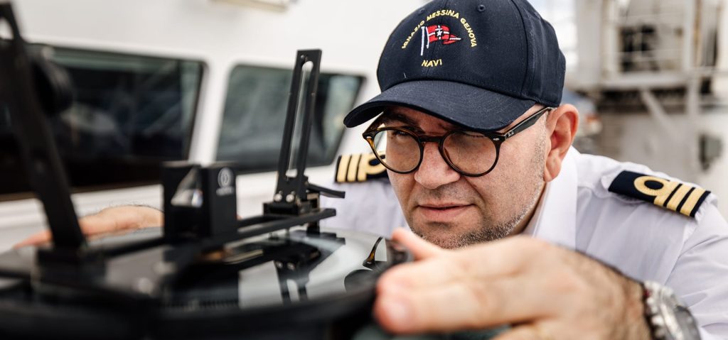 Ship Captain in uniform using a bearing repeater on the bridge wing.