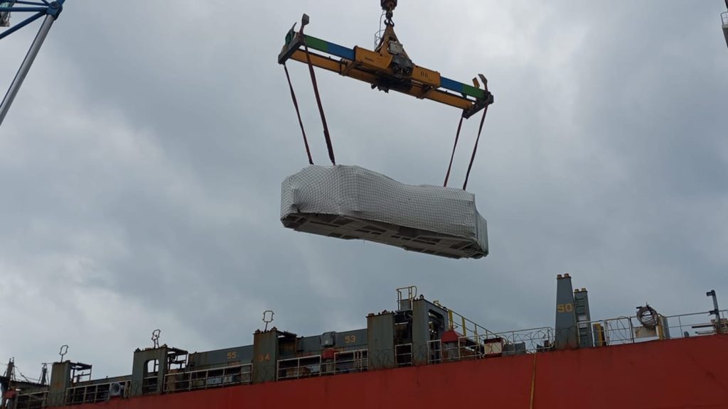 Heavy machinery wrapped in protective white film being lifted onto a container ship.