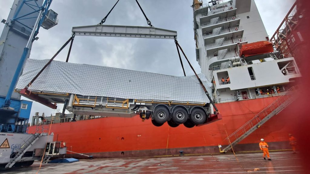A heavy-duty trailer being hoisted by a crane next to the hull of the Jolly Rosa.