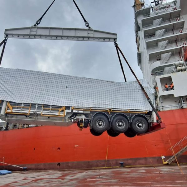 A heavy-duty trailer being hoisted by a crane next to the hull of the Jolly Rosa.
