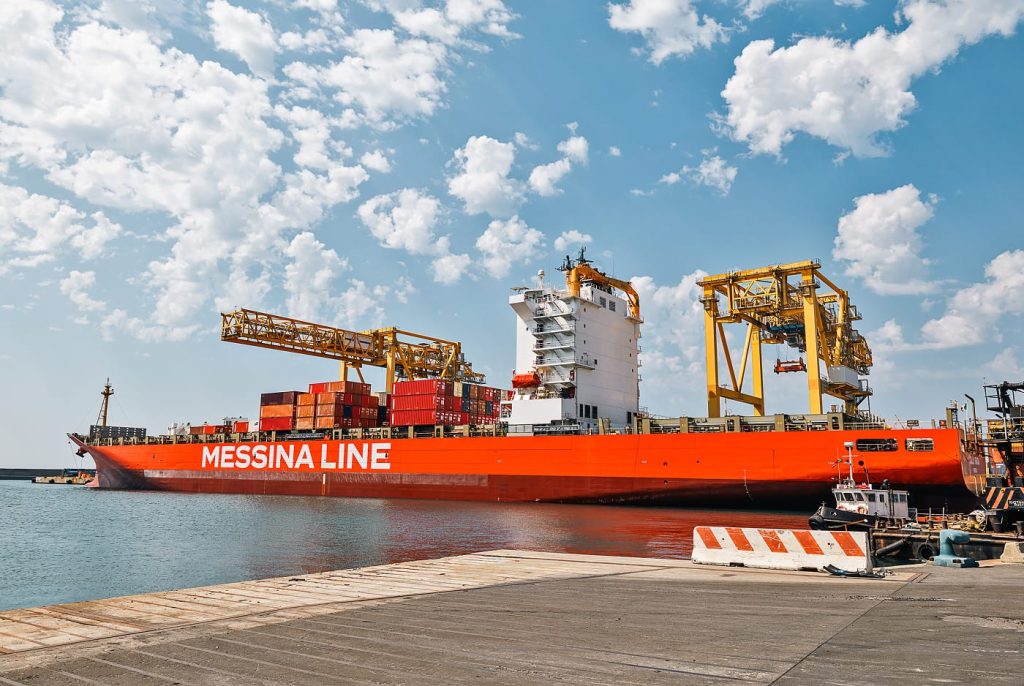 Side profile of the Jolly Giada container ship docked under large yellow port cranes.