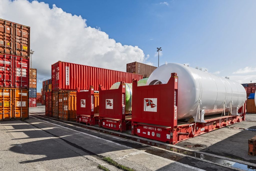 A massive white pressurized tank unitized on a flatrack in a port storage area.