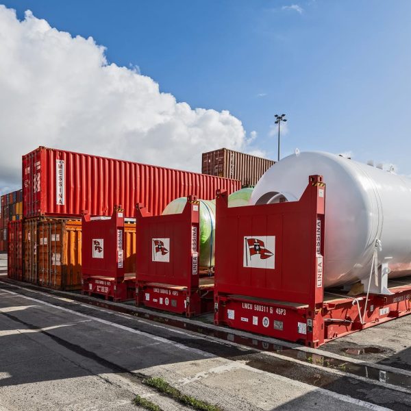 A massive white pressurized tank unitized on a flatrack in a port storage area.